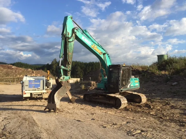 Green tracked excavator with a bucket and thumb attachment on an earthworks site, with a field service truck parked beside it