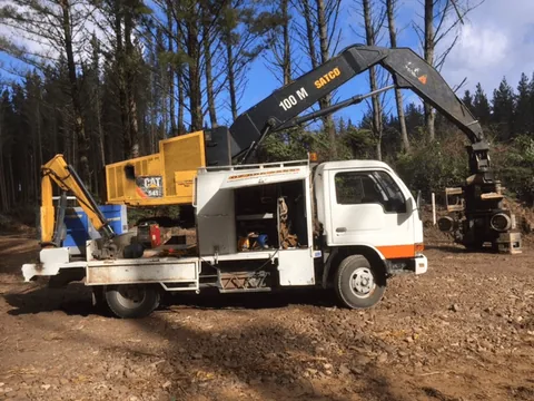 White field service truck with a yellow CAT unit and tool lockers on its deck, parked beside a Satco forestry machine on a cleared site among pine trees
