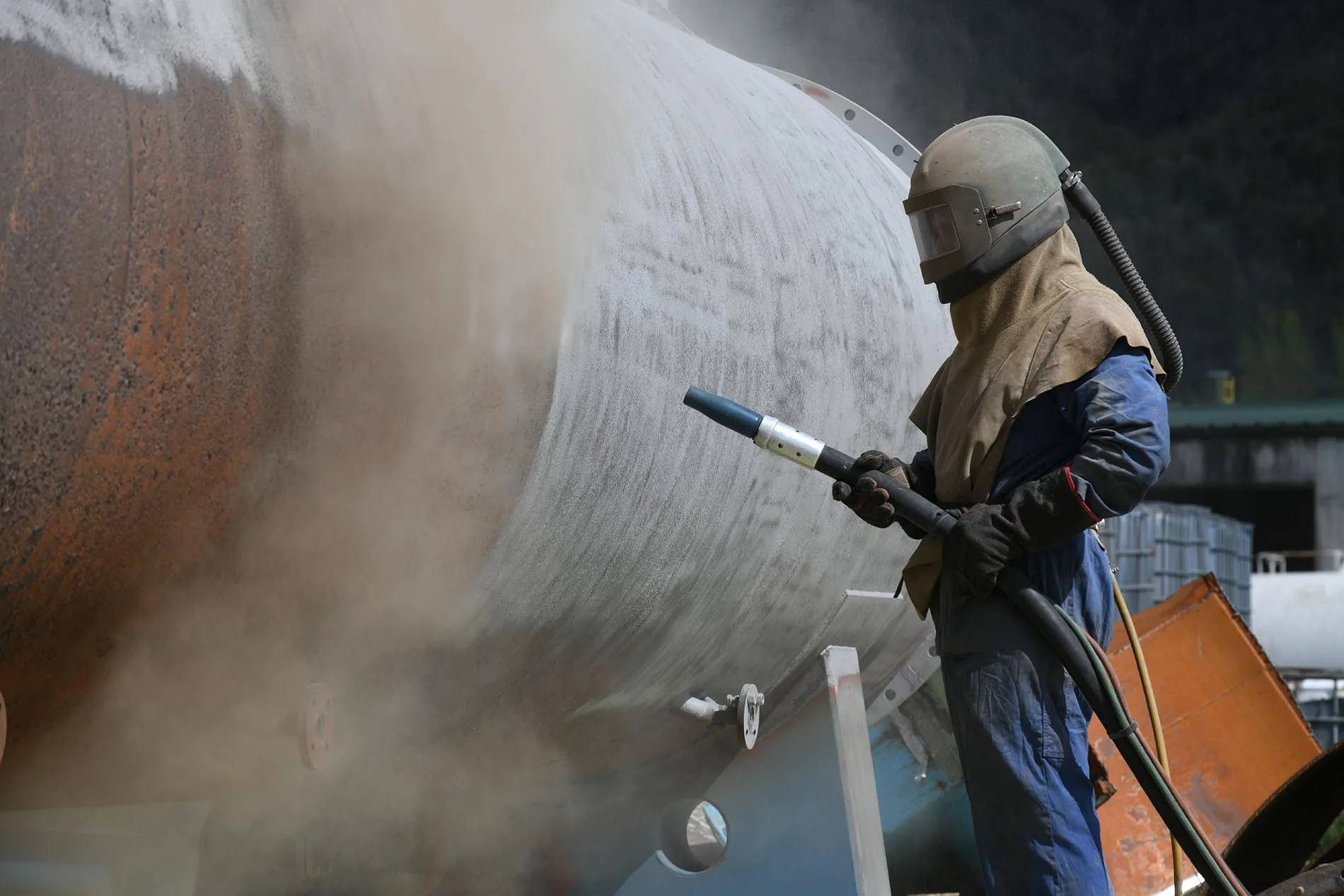 Stock photograph of an operator in a blast helmet and hood sandblasting rust off a large steel tank in a cloud of dust